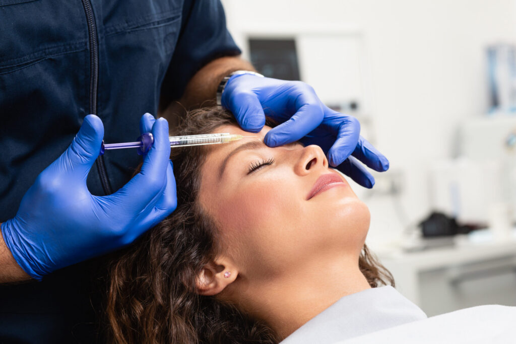 Close-up of a healthcare provider giving a facial injection to a woman with eyes closed, using a syringe and blue gloves.