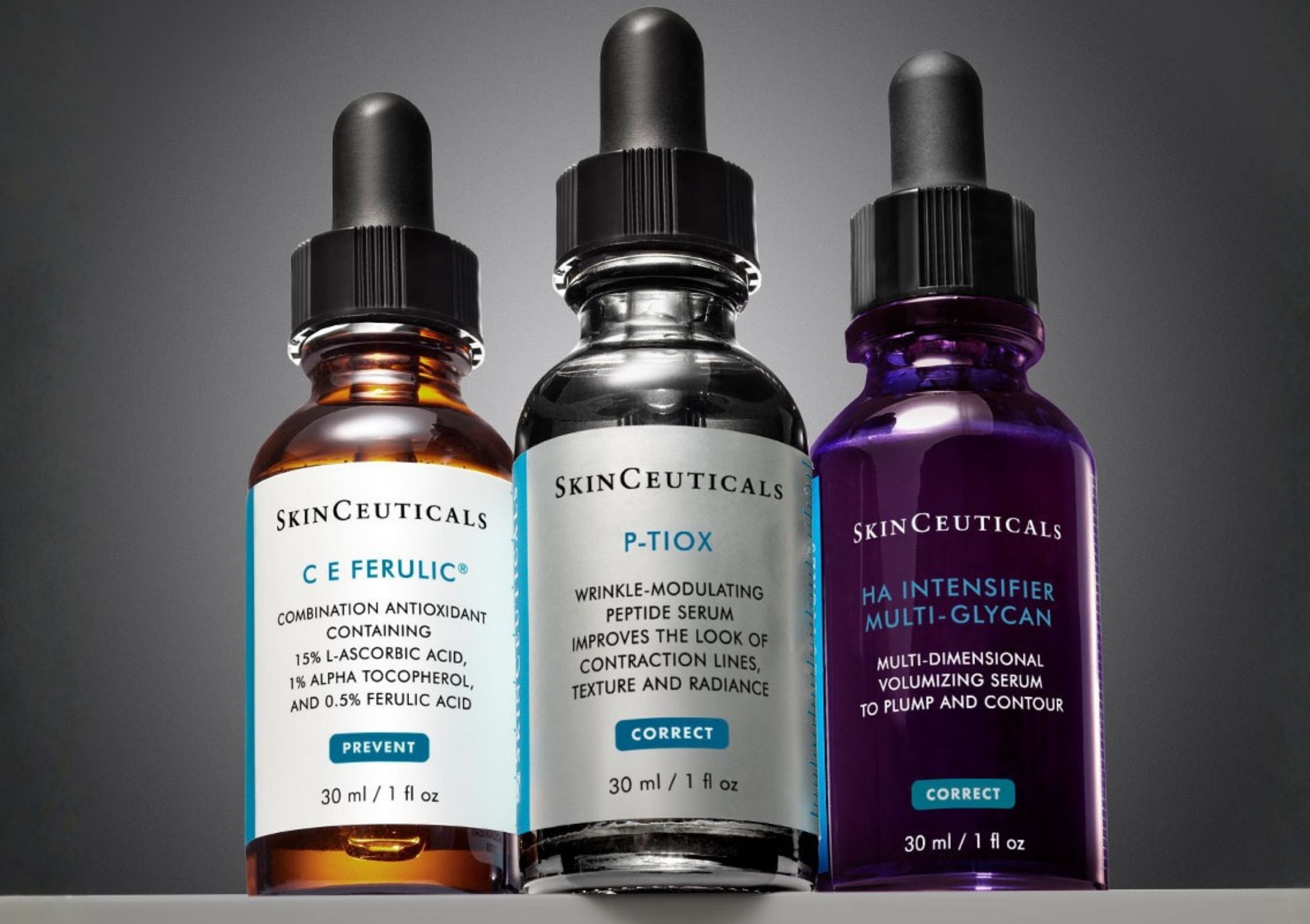 Three SkinCeuticals dropper bottles in a row: orange, clear, and purple liquids with black droppers against a gray background.