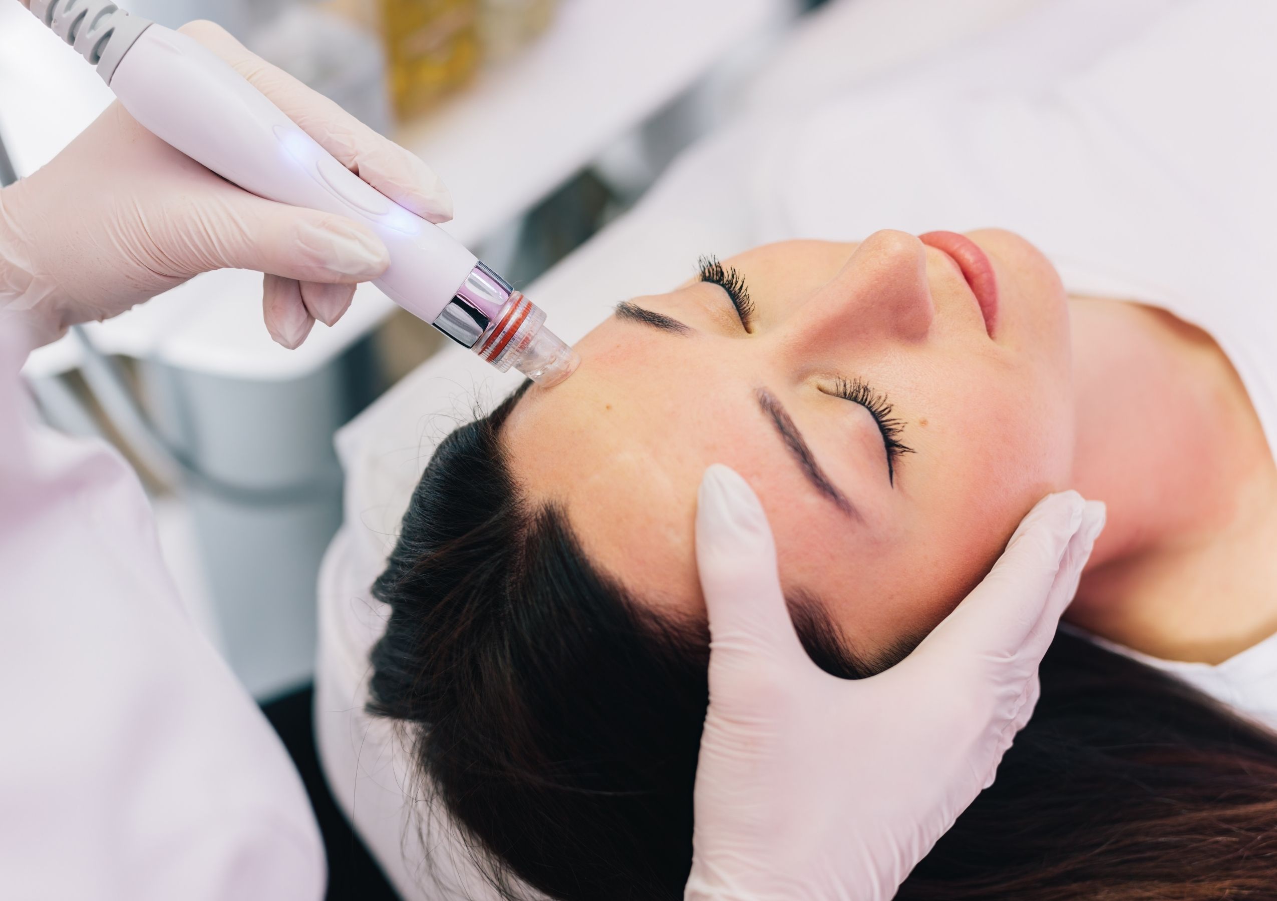 Close-up of a gloved technician using a handheld cosmetic device near a client's eye as the person lies resting.